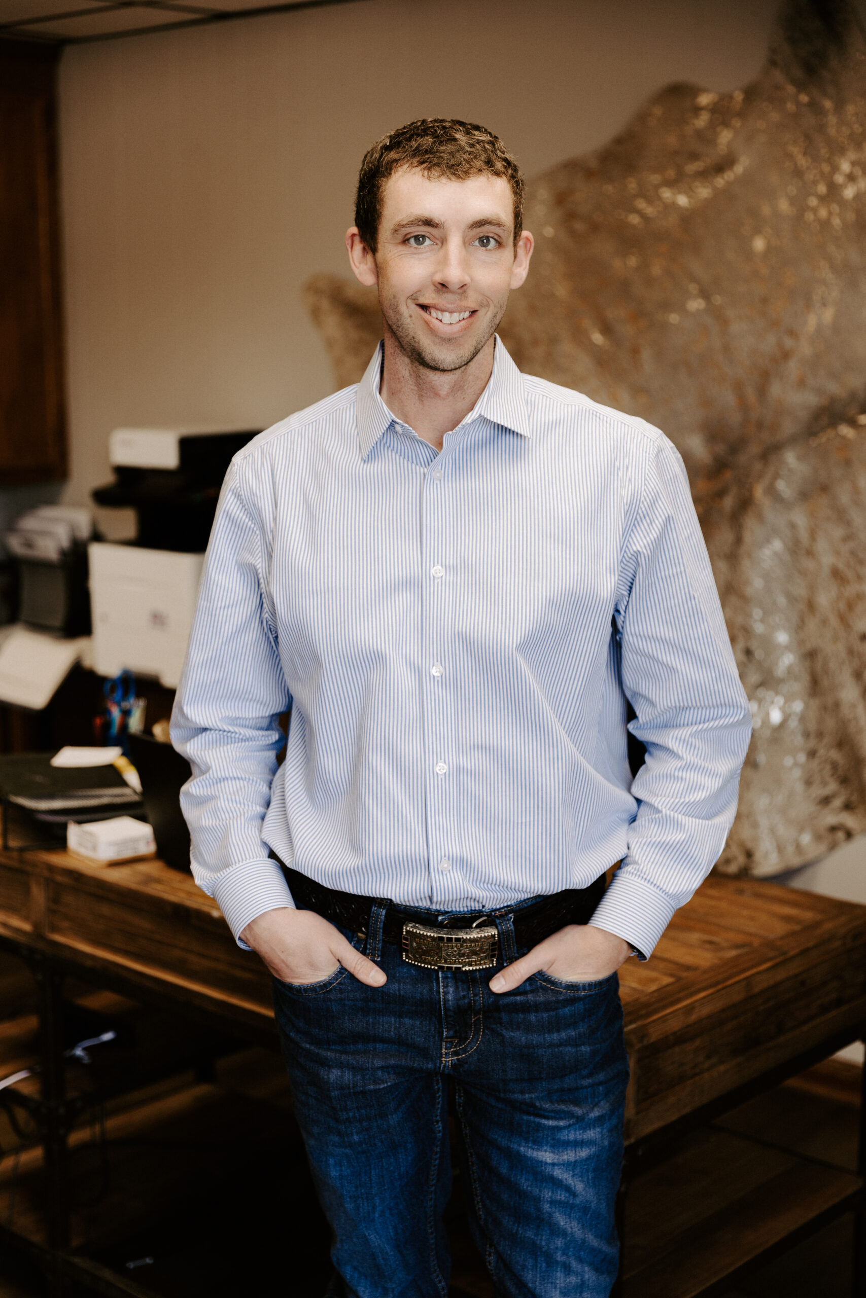 Professional man in a blue striped shirt and jeans, standing confidently in an office setting with a wooden table and office supplies in the background.