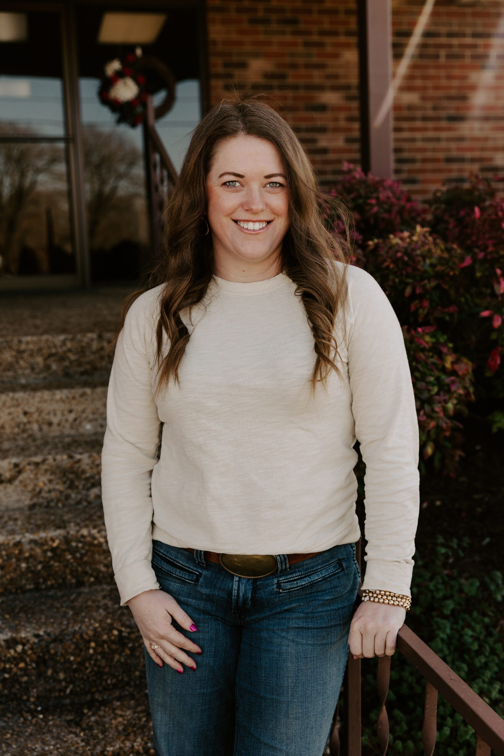 Smiling woman in a beige long-sleeve shirt and blue jeans standing on outdoor steps, with a decorative wreath in the background and colorful plants nearby.