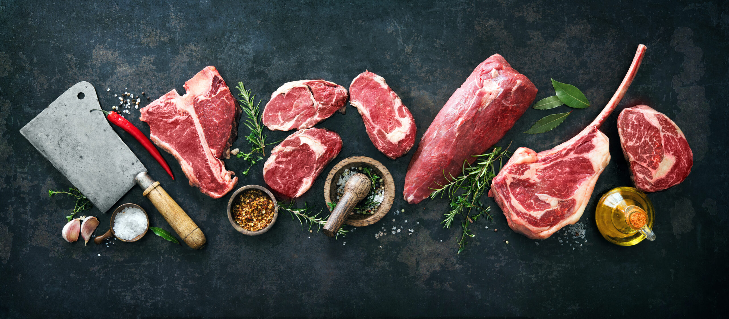 Assorted cuts of fresh red meat arranged on a dark surface, featuring a cleaver, spices, herbs, and garlic, highlighting culinary preparation and gourmet cooking.