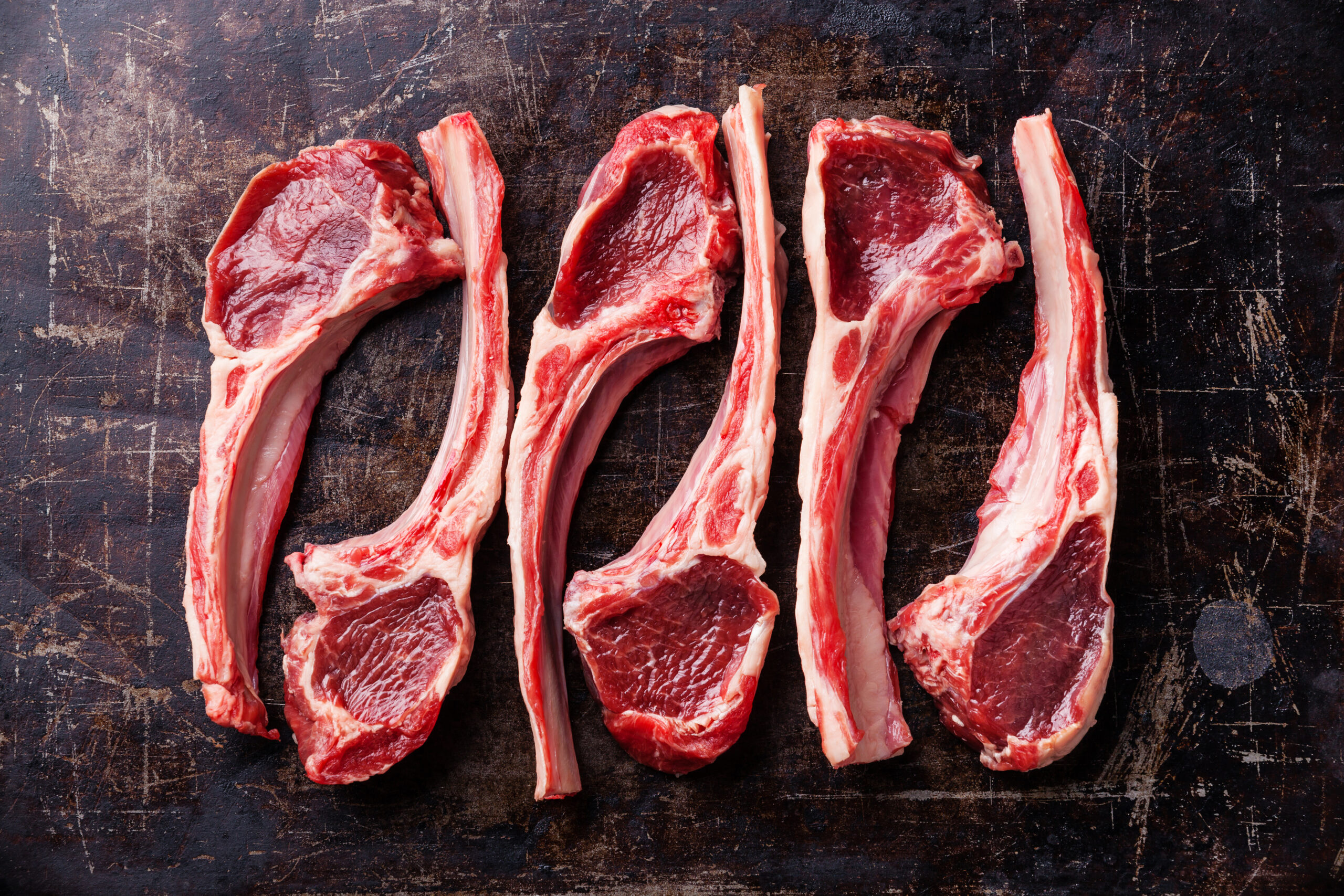 Four raw lamb chops arranged on a rustic wooden surface, showcasing their marbled red and white meat. Ideal for grilling or roasting, these cuts highlight the quality and freshness of lamb.