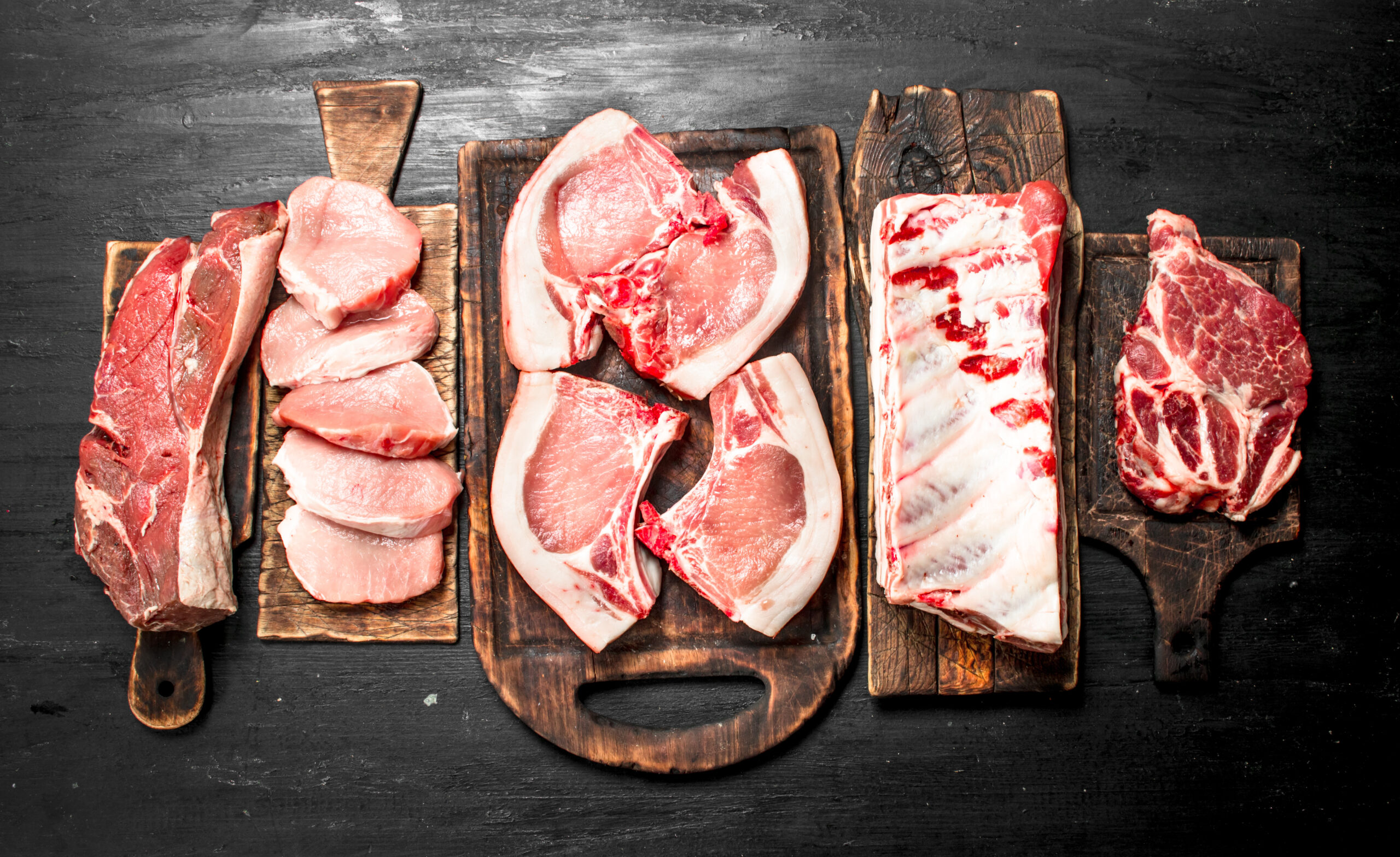 Variety of fresh raw meats displayed on wooden cutting boards, including pork chops, pork belly, and beef cuts, arranged on a dark surface, highlighting their rich textures and colors for culinary inspiration.